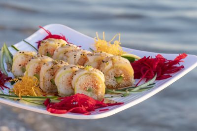 "A fresh local fish sushi roll artfully plated on an outdoor table with a scenic beach and ocean background."